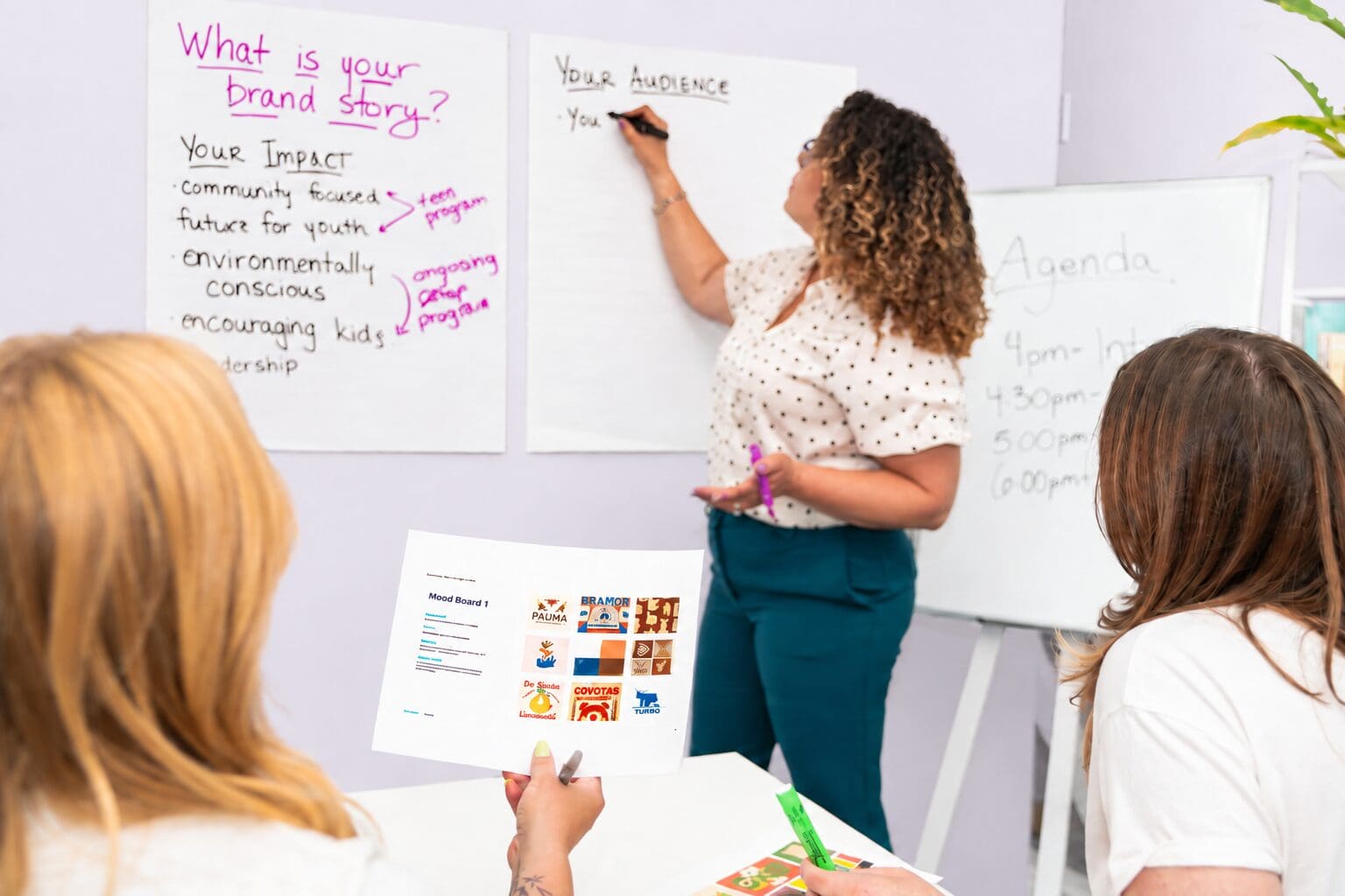 Three women are in a bright room. One writes on a paper about Your Audience, while another holds a branding handout. Notes about a brand story and agenda are on whiteboards. The mood is collaborative and focused.