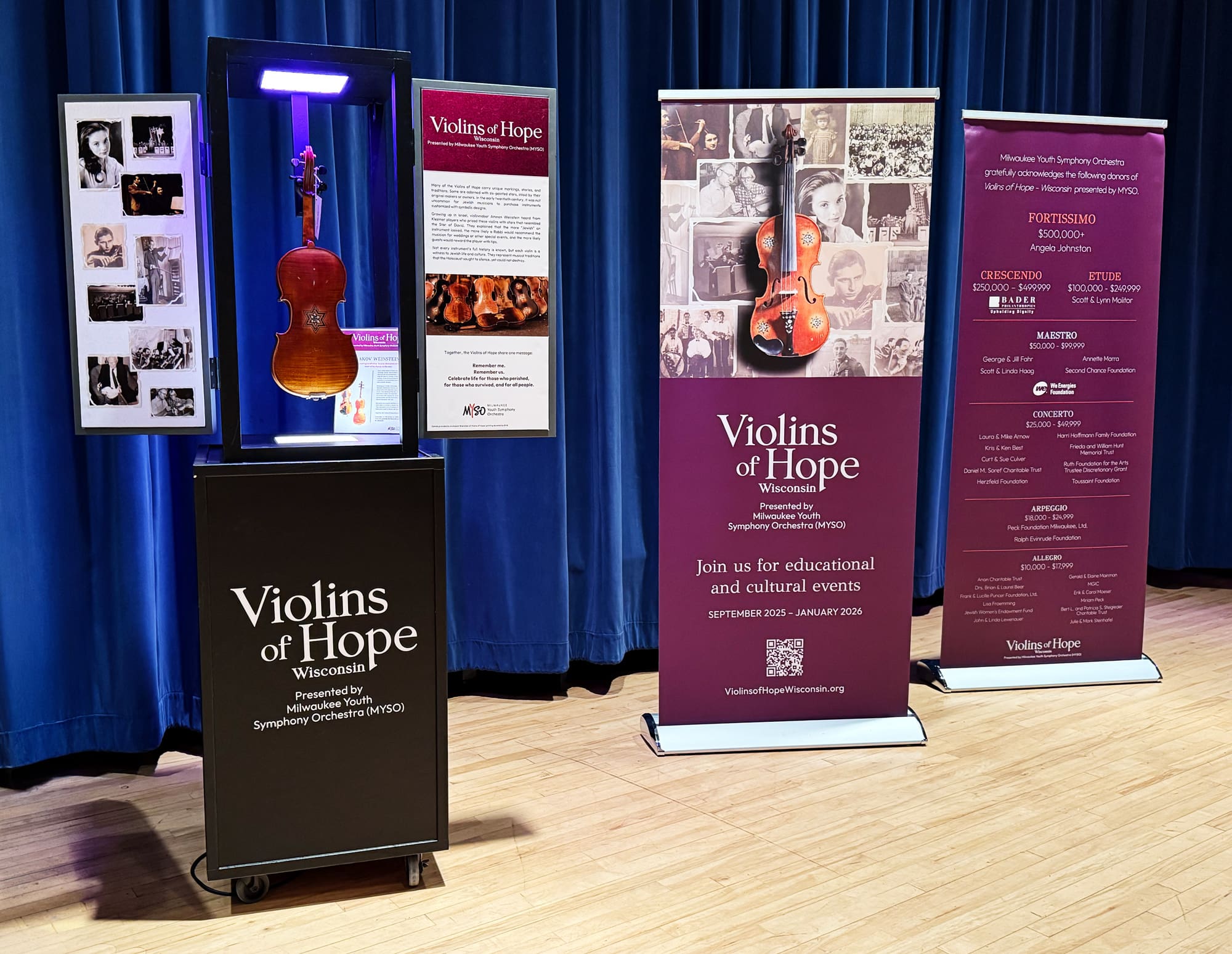A violin on display in a glass case stands between two informational banners for Violins of Hope, an educational and cultural event series, with a collage of black-and-white photos on the left.