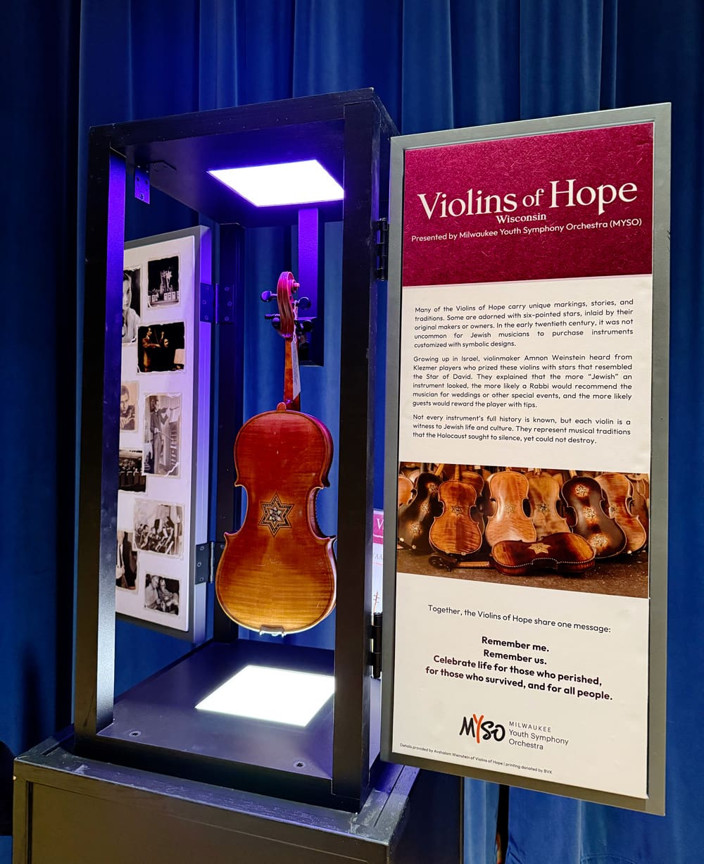 A violin is displayed in a glass case next to an informational sign titled Violins of Hope, with photos and text about the exhibit by Milwaukee Youth Symphony Orchestra on a blue backdrop.