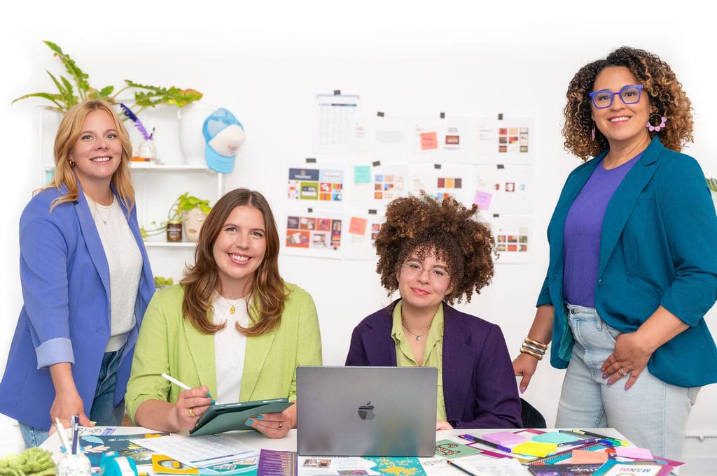 Four women in colorful blazers work together at a desk with laptops, papers, and office supplies. A vision board and plants are visible in the brightly lit, creative workspace.