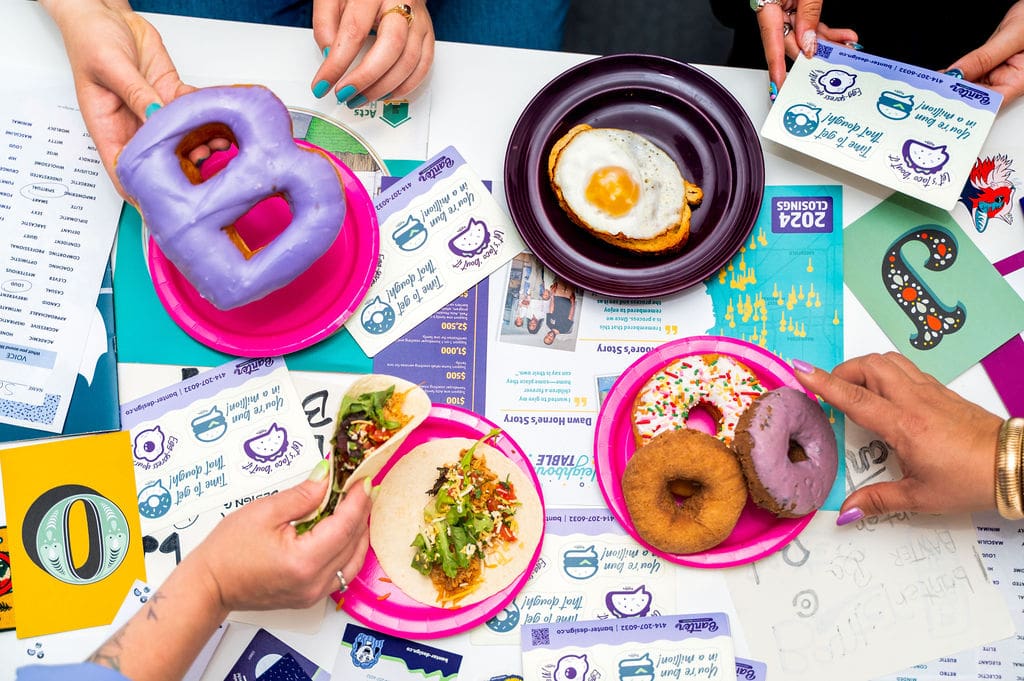 Four people share colorful donuts, a taco, and a plate with a sunny-side-up egg over papers, stickers, and infographics on a table with a playful, creative atmosphere.