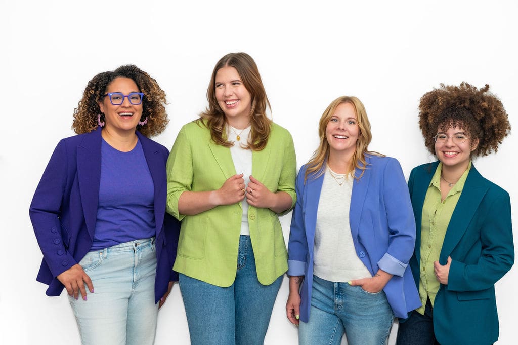 Four women stand together, smiling and wearing colorful blazers and jeans against a plain white background.