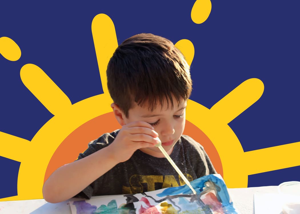 A young boy paints with watercolors at a table, concentrating on his artwork. A bright, cartoon sun with yellow rays is illustrated behind him on a blue background.