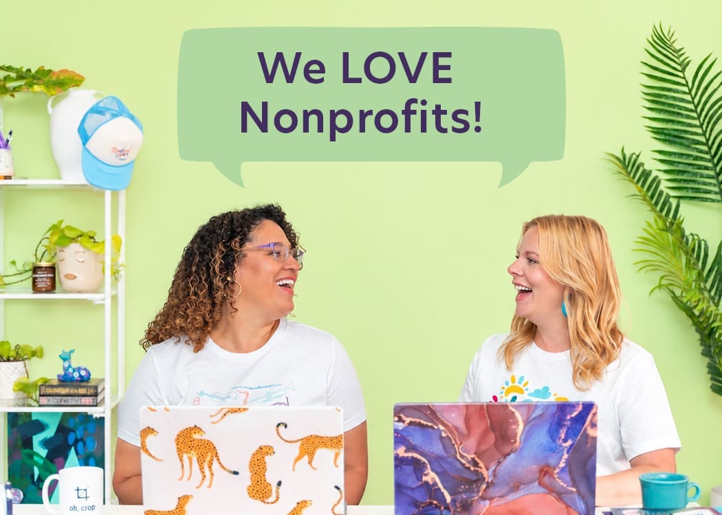 Two women sit at a colorful desk with laptops, smiling at each other. A speech bubble above them says, We LOVE Nonprofits! The background is green with plants and office decor.
