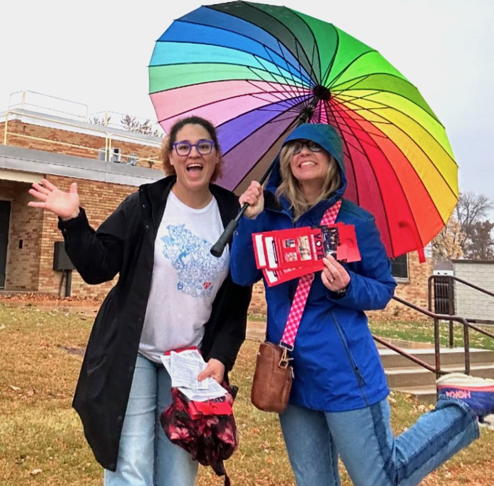 Two women stand outside under a large rainbow umbrella, one waving and the other holding red information pamphlets.